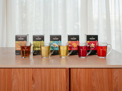 Heimbs Teepyramiden mit Glas Fünf Tee-Pyramidenpackungen im Hintergrund. Davor stehen fünf gefüllte Teegläser auf einem Holztisch.