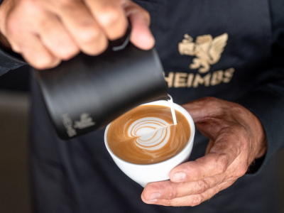 Heimbs Latte Art Cappuccino Barista gießt mit Milchkännchen Herz in die Cappuccino Tasse. Heimbs Logo im Hintergrund
