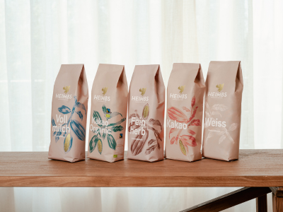 Heimbs Trinkschokolade Vollmilch, Feinherb, Weisse Fünf Trinkschokoladen Beutel von der Seite auf einem braunen Holztisch